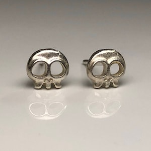 May include: A pair of silver skull stud earrings. The skulls are small and detailed, with hollow eye sockets and a small nose.