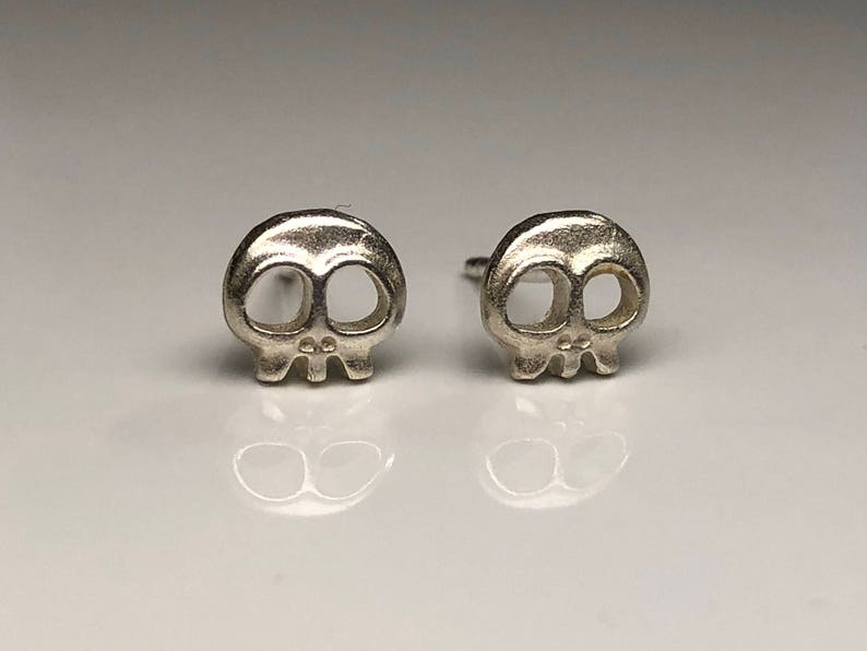 May include: A pair of silver skull stud earrings. The skulls are small and detailed, with hollow eye sockets and a small nose.