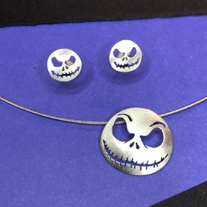 May include: A silver necklace and earrings set featuring a cutout design of Jack Skellington's face from the movie The Nightmare Before Christmas. The necklace has a silver chain and a larger pendant with the face design. The earrings are smaller and feature the same design.