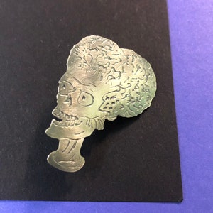 Pode incluir: Pin de metal prateado com um perfil detalhado de uma cabe&ccedil;a alien&iacute;gena com um design texturizado que se assemelha a um c&eacute;rebro.