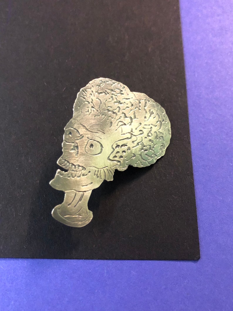 Pode incluir: Pin de metal prateado com um perfil detalhado de uma cabe&ccedil;a alien&iacute;gena com um design texturizado que se assemelha a um c&eacute;rebro.