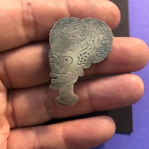 Pode incluir: Pin de metal prateado com um perfil detalhado de um alien&iacute;gena fict&iacute;cio com uma cabe&ccedil;a grande e um design texturizado semelhante a um c&eacute;rebro.