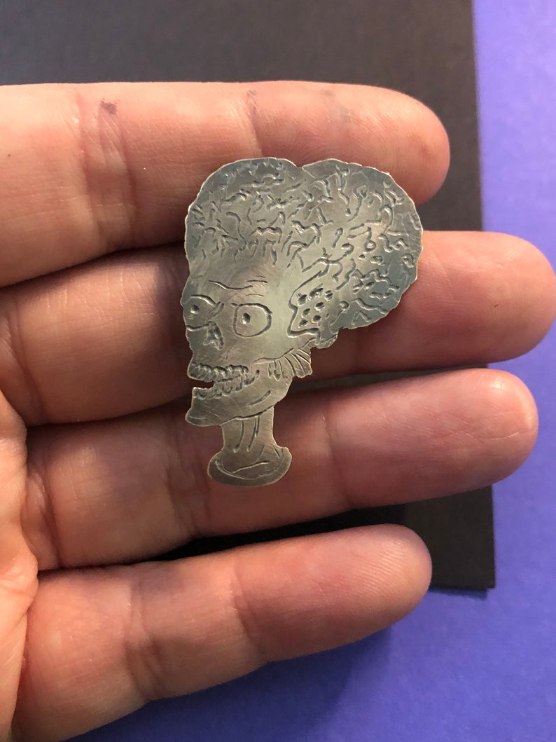 Pode incluir: Pin de metal prateado com um perfil detalhado de um alien&iacute;gena fict&iacute;cio com uma cabe&ccedil;a grande e um design texturizado semelhante a um c&eacute;rebro.