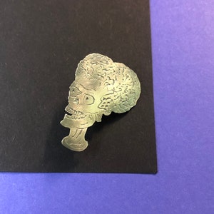 Pode incluir: Pin de metal prateado com um perfil detalhado de um alien&iacute;gena de desenho animado com uma cabe&ccedil;a grande e um corpo pequeno. O alien&iacute;gena tem uma cabe&ccedil;a grande e redonda com uma superf&iacute;cie texturizada e um corpo pequeno e fino com um pesco&ccedil;o longo.