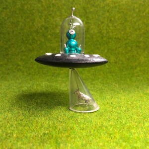 Peut inclure: Un OVNI miniature avec une figurine extraterrestre turquoise à l'intérieur, posé sur un support en plastique transparent avec une figurine d'animal argentée en dessous. L'OVNI a une base noire avec des points blancs.