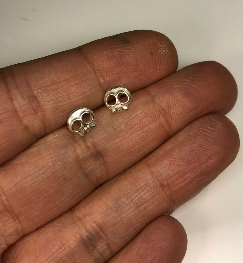 May include: A pair of silver skull stud earrings. The earrings are small and delicate, and they have a simple, minimalist design.