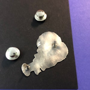 Pode incluir: Um alfinete de metal prateado em forma de ilha com uma superf&iacute;cie texturizada. O alfinete tem uma parte de tr&aacute;s de metal prateado com um poste de alfinete.