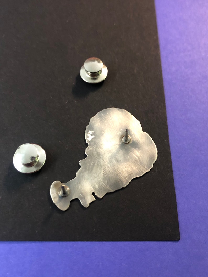 Pode incluir: Um alfinete de metal prateado em forma de ilha com uma superf&iacute;cie texturizada. O alfinete tem uma parte de tr&aacute;s de metal prateado com um poste de alfinete.