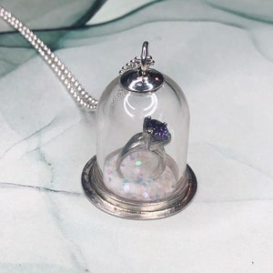 Könnte beinhalten: Eine silberne Halskette mit einem Glaskuppel-Anhänger, der einen silbernen Ring mit einem violetten Edelstein und schimmernden Glitzer enthält.