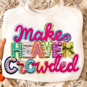 May include: White sweatshirt with the phrase "Make Heaven Crowded" in colorful, appliqued letters. The letters are embellished with pearls, ribbons, and patterned fabrics. The sweatshirt has a crew neck.