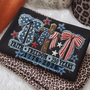 May include: Black fabric with an embroidered design featuring a cross, bows, and stars in red, white, and blue. The text reads "FAITH FREEDOM 1776-2026 250 YEARS". The fabric is resting on a leopard print fabric.