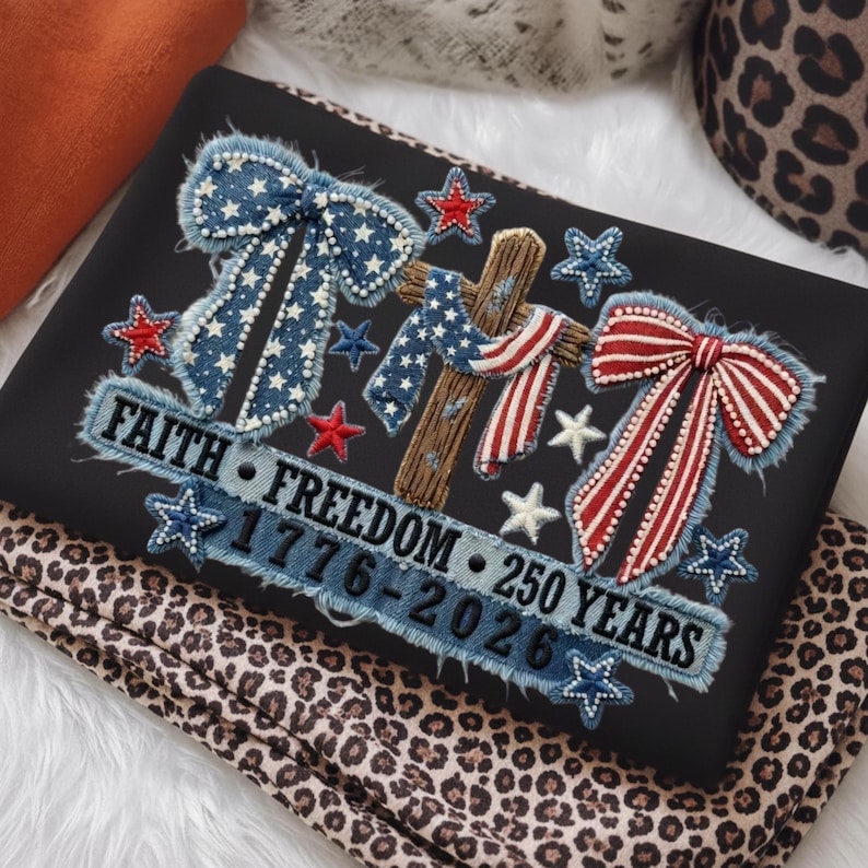 May include: Black fabric with an embroidered design featuring a cross, bows, and stars in red, white, and blue. The text reads "FAITH FREEDOM 1776-2026 250 YEARS". The fabric is resting on a leopard print fabric.