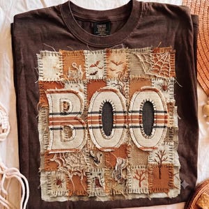 Può includere: Una t-shirt marrone scuro con una grande grafica rettangolare in stile patchwork. Il design mostra la parola BOO in grandi lettere di tessuto color crema con strisce arancioni e nere, circondate da quadrati cuciti con ragnatele, pipistrelli e alberi.