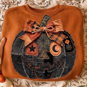 Op de afbeelding: Een roestoranje sweater met ronde hals en een grote, centrale pompoenapplicatie. De pompoen is gemaakt van zwarte corduroy en denim patches met gerafelde randen, een geruite stoffen strik en geborduurde details zoals een ster en maan.