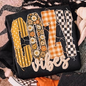 May include: A black sweatshirt featuring the word COZY in large, frayed-edge fabric letters with varied patterns including yellow polka dots, floral prints, orange gingham, and black-and-white checkers, with the word vibes in white pearls.