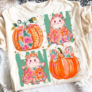 Puede incluir: Una camiseta de color crema con un estampado gráfico de dos calabazas naranjas y dos fantasmas decorados con patrones florales rosas y naranjas. El diseño incluye acentos de pinceladas verticales en color verde azulado y puntos dorados.