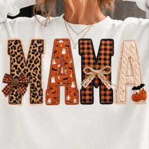 May include: A white sweatshirt featuring the word MAMA in large block letters. Each letter has a unique pattern: leopard print with a plaid bow, orange with ghosts and bats, black and orange plaid with a twine bow, and cream cable knit with pumpkins.