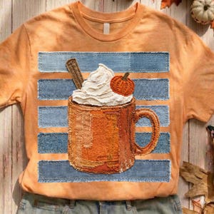 Op de afbeelding: Een perzikkleurig t-shirt met een afbeelding van een oranje mok gevuld met slagroom, een kaneelstokje en een kleine pompoen. Het ontwerp is geplaatst over horizontale blauwe denim-gestreepte banen met gerafelde randen.