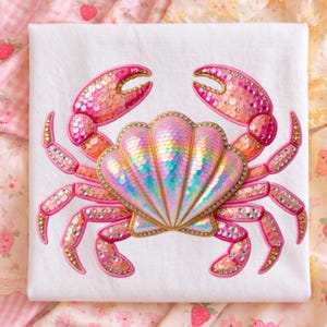 May include: A white background features a decorative crab design. The crab has pink and orange sequined claws and legs, and a shimmering, iridescent shell. Rhinestones and gold trim embellish the design, creating a glamorous, coastal theme.