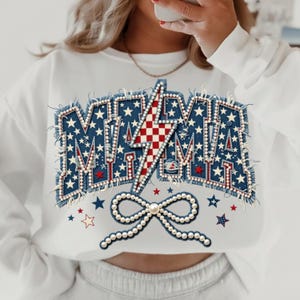 Op de afbeelding: Witte sweatshirt met een denim-stijl "MAMA"-opdruk. De letters zijn versierd met sterren en een rood-witte bliksemschicht. Een parelboog en verspreide sterren maken het ontwerp compleet.