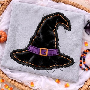 Op de afbeelding: Een lichtgrijs t-shirt met een grote applicatie van zwart fluweel in de vorm van een heksenhoed met gerafelde randen. De hoed is voorzien van een paarse band, een oranje gesp en oranje decoratieve stiksels in het midden van het shirt.