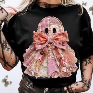 Può includere: Una t-shirt nera con un'applicazione a forma di fantasma realizzata con vari ritagli di tessuto rosa e crema, tra cui pizzo, tessuto scozzese e velluto a coste. Il fantasma è decorato con un grande fiocco di nastro rosa sfilacciato e ciondoli.