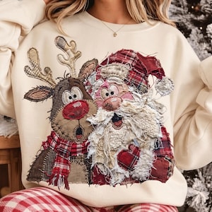 May include: A cream-colored crewneck sweatshirt featuring a large, textured applique design of a reindeer and Santa Claus. The reindeer has a red nose and plaid scarf, while Santa wears a red plaid hat and coat with frayed fabric edges.