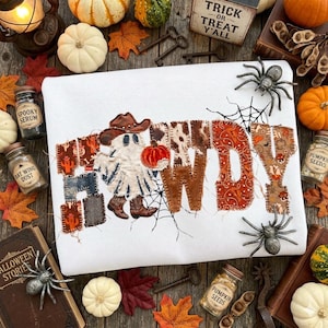 May include: A white sweatshirt featuring the word HOWDY appliqued in various patterned fabrics. The letter O is replaced by a ghost wearing a cowboy hat and boots, holding a pumpkin. The design includes spider web embroidery and frayed edges.