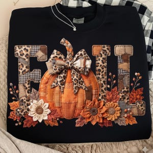 May include: A black crewneck sweatshirt featuring the word FALL in large, patchwork-style letters with leopard and houndstooth patterns. An orange pumpkin with a leopard print bow sits in the center, surrounded by fabric flowers and autumn leaves.