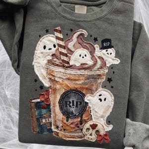 May include: A charcoal gray sweatshirt featuring a fabric applique of a coffee cup topped with whipped cream and ghosts. The cup has a circular patch with the text R.I.P. and is surrounded by ghosts, a book labeled R.I.P., and a skull with a red bow.