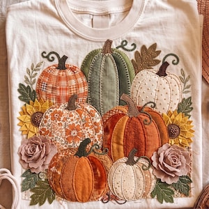 May include: A cream-coloured t-shirt featuring a central appliqué design of various pumpkins in shades of orange, green, and white. The pumpkins are accented with fabric sunflowers, dusty rose fabric flowers, and green fabric leaves.