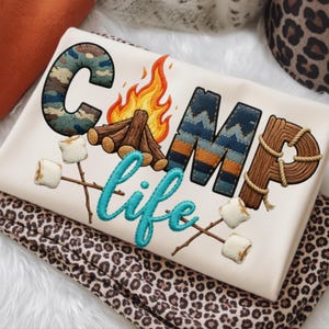 May include: Cream sweatshirt featuring the embroidered text "CAMP life". The word "CAMP" is designed with camouflage, blue and orange patterns, and wood textures. The design includes a campfire, flames, and marshmallows on sticks.
