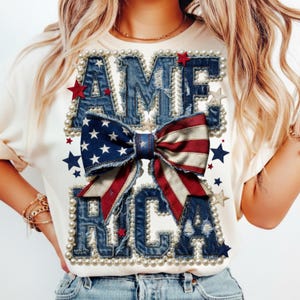 Pode incluir: Camiseta creme com a palavra "AMERICA" em letras de ganga, contornada com pérolas. Uma fita com as cores da bandeira americana está centrada abaixo do "A". Estrelas vermelhas e azuis estão espalhadas pelo design.