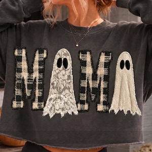 May include: A charcoal gray long-sleeve sweatshirt featuring the word MAMA in large applique letters. The M letters are made of black and white buffalo plaid fabric, while the A letters are shaped like ghosts with lace and textured fabric.