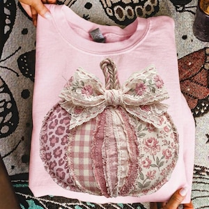 May include: A light pink crewneck sweatshirt featuring a large, central pumpkin applique. The pumpkin is constructed from various fabric patches, including leopard print, pink gingham, and floral patterns, topped with a decorative lace bow.