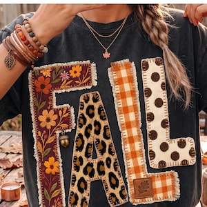 May include: A black t-shirt featuring the word FALL in large, textured letters. The F is dark purple with floral embroidery and an acorn charm. The A has a leopard print pattern. The Ls feature orange gingham and cream with brown polka dots.
