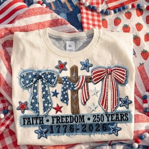 May include: Cream-colored XL t-shirt with patriotic embroidery. Features a cross draped with a flag, bows, and stars in red, white, and blue. Text reads "FAITH FREEDOM 250 YEARS 1776-2026".