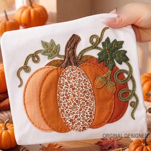 May include: A white fabric square featuring a central pumpkin appliqué. The pumpkin is composed of orange and rust-coloured fabric segments, with one central section displaying a small floral pattern. It includes a brown stem and green vines.