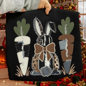 May include: Black cushion featuring a bunny with a leopard print design and a brown bow. Two carrots flank the bunny, with green tops and denim-like orange bodies. The cushion is held up by a person.