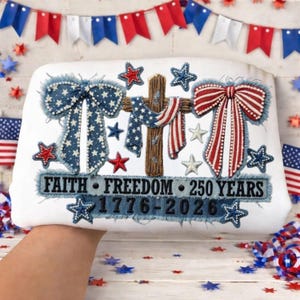 May include: White rectangular pillow with patriotic embroidery. Features blue and white star bows, a cross draped with a flag, and red and white striped bow. Text reads "FAITH FREEDOM 250 YEARS 1776-2026". Decorative stars complete the design.