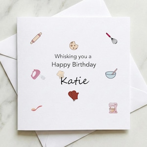 May include: A square white birthday card with the message "Whisking you a Happy Birthday Katie" and baking-themed illustrations. Includes a rolling pin, whisk, and mixer. The card is inside a white envelope.