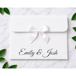 May include: A white rectangular envelope with a textured finish, featuring a centred white satin ribbon bow on the flap. The front of the envelope displays the black printed text Emily & Josh in a cursive script font, centred below the ribbon.