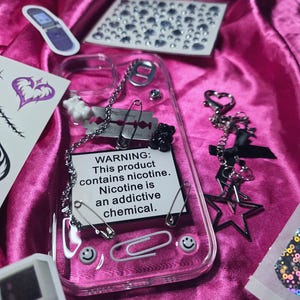 Op de afbeelding: Transparante telefoonhoes met ketting, veiligheidsspelden en een scheermesje bedel. De hoes heeft een waarschuwingslabel: "Dit product bevat nicotine. Nicotine is een verslavende chemische stof." Stickers en een zwarte sterrenbedel.