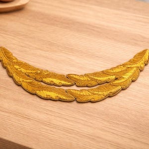 Puede incluir: Dos apliques de plumas bordadas en color dorado, dispuestas en forma curva. Los apliques presentan costuras detalladas y están colocados sobre una superficie de madera de color marrón claro. Las plumas son un artículo de artesanía decorativa.