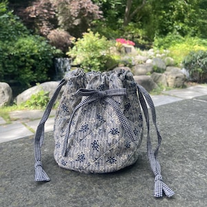 Può includere: Una borsa a sacchetto trapuntata con coulisse, caratterizzata da un motivo floreale blu scuro e bianco su sfondo a righe. La borsa presenta un'apertura arricciata chiusa da un nastro a quadretti vichy blu scuro e bianco che pende ai lati.