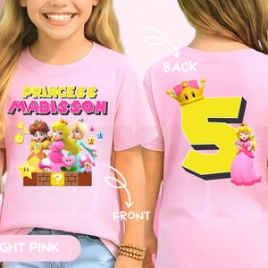 May include: A light pink short-sleeved t-shirt featuring a front graphic of Princess Peach, Daisy, and Rosalina with the text Princess Madisson. The back of the shirt displays a large yellow number five topped with a crown and Princess Peach.