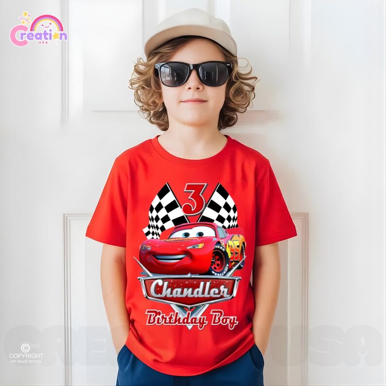 May include: A red t-shirt featuring a graphic of the character Lightning McQueen from the movie Cars, with the number 3, checkered flags, and the text "Chandler Birthday Boy". The shirt is worn by a person wearing sunglasses and a cap.