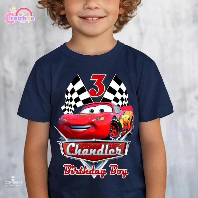 May include: Navy blue t-shirt featuring a red Lightning McQueen race car graphic with the number 95. The shirt also includes the name "Chandler" and the text "Birthday Boy" in a red and white design, with a number 3 and checkered flags.