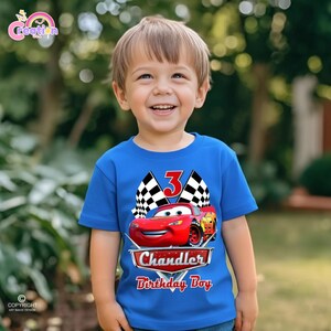 May include: A blue t-shirt featuring a red race car graphic with the number 3, checkered flags, and the text "Chandler Birthday Boy." The shirt is worn by a young child.