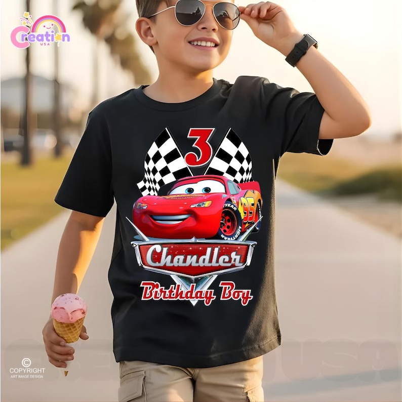 May include: Black t-shirt featuring a red race car graphic with the number 3, checkered flags, and the text "Chandler Birthday Boy." The shirt is worn by a person holding an ice cream cone.